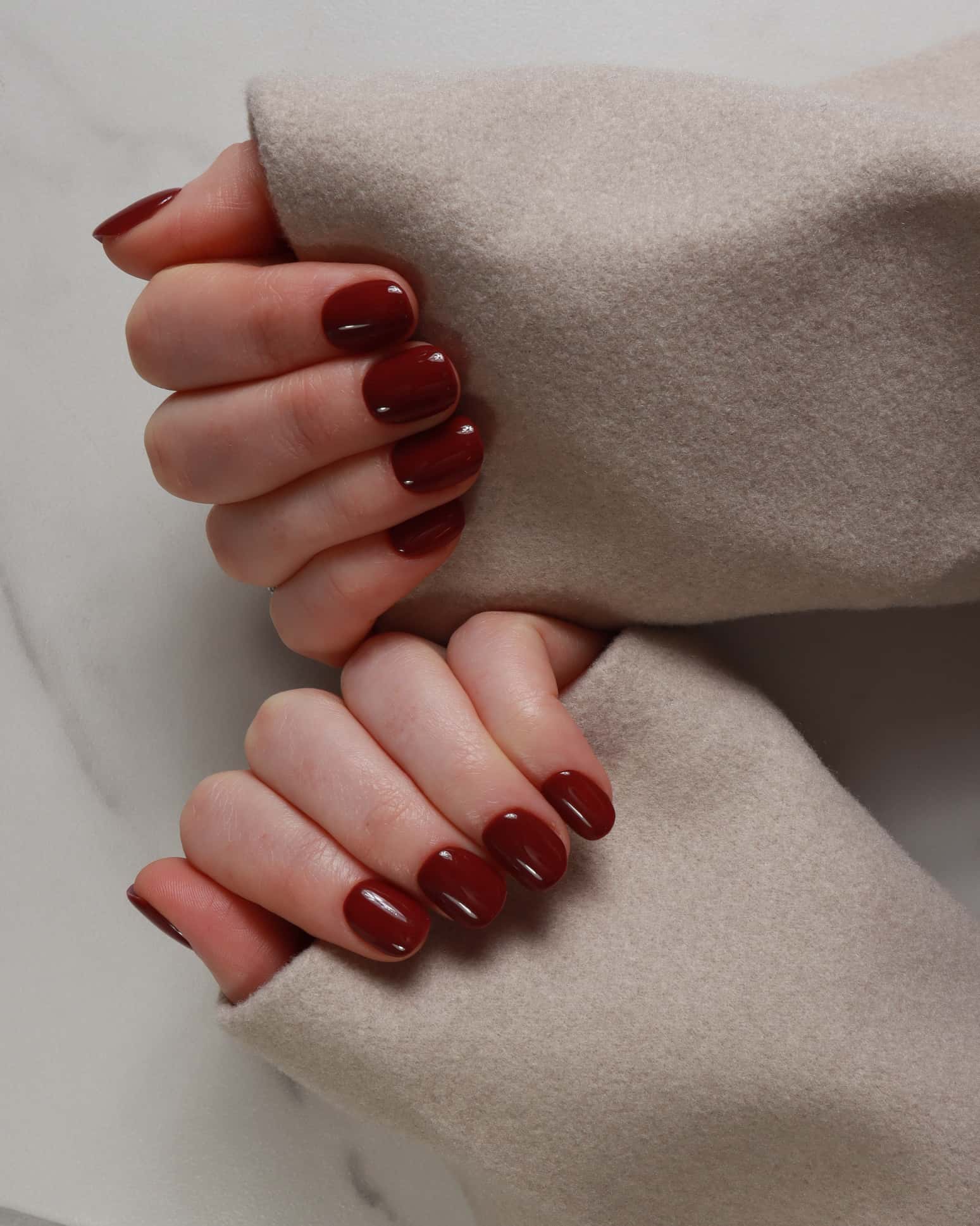 Close-up of hands with dark red nail polish wearing a beige coat.