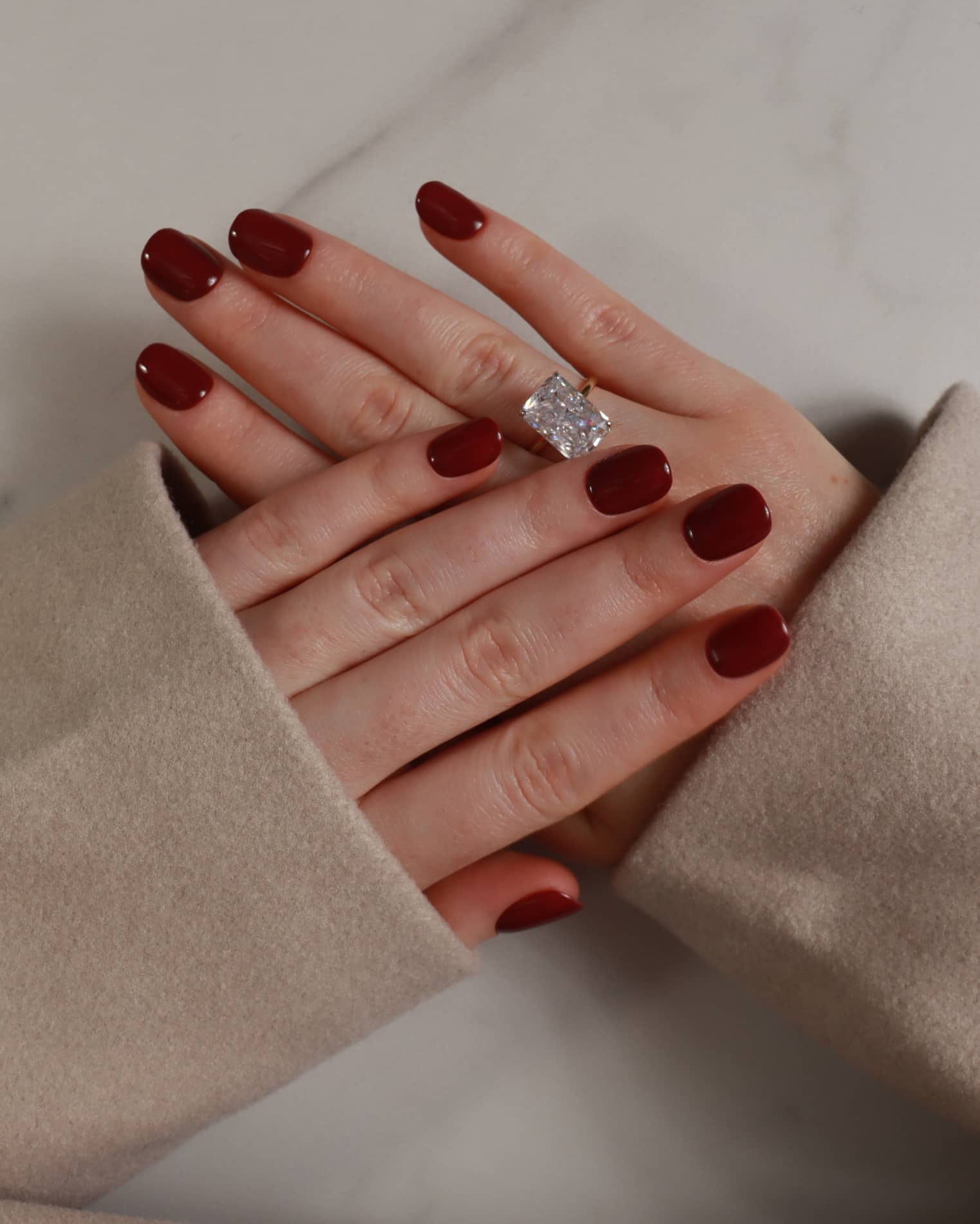 Hand with red nail polish wearing a diamond ring, wearing beige gloves on a neutral background